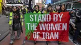 Vrouwen lopen over straat met een spandoek. Op het spandoek staat de tekst 'Stand by the women of Iran'.