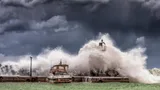 Foto van storm waarbij grote golf vuurtoren en klein bootje overspoelt.
