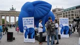 Foto van protestanten en een grote, blauwe opblaasolifant voor de Brandenburger Tor in Berlijn. Op de grond protestborden met daarop: It's time wealth gets taxed the same as work!