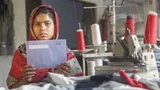 A woman wearing a headscarf sits next to a sowing machine. She holds a tax envelope.