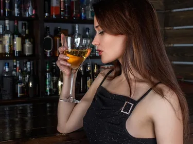 Een vrouw houdt een glas wijn voor haar gezicht. Op de achtergrond een bar.