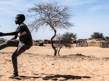 Een Afrikaanse jongen houdt een bal hoog. Hij staat op een dorre vlakte met hutten op de achtergrond.