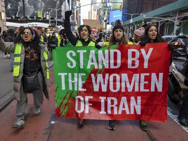Vrouwen lopen over straat met een spandoek. Op het spandoek staat de tekst 'Stand by the women of Iran'.
