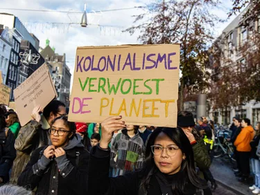 Vrouw in protestmars houdt bord omhoog met daarop de tekst: Kolonialisme verwoest de planeet