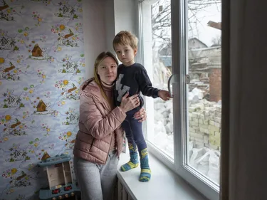 Ana houdt haar zoon vast, terwijl hij op de vensterbank van een slaapkamer staat. Buiten ligt een stapel stenen.
