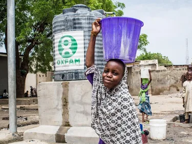 Een meisje vult een emmer bij een tank met het Oxfam logo. Ze houdt de emmer op haar hoofd.