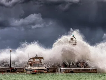 Foto van storm waarbij grote golf vuurtoren en klein bootje overspoelt.