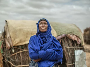 Een Afrikaanse vrouw met een hoofddoek staat voor een hut.