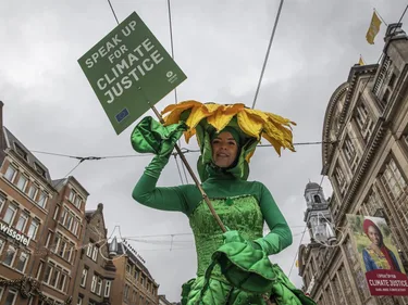 Een vrouw verkleed als bloem houdt een bord vast met de tekst 'Speak up for climate justice'.