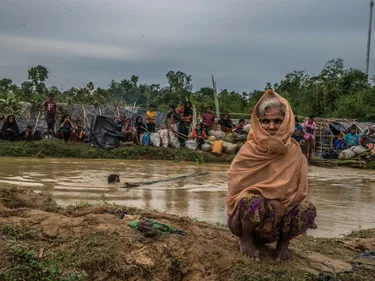 Een oude vrouw voor een rivier met vies water. Aan de andere kant van de rivier zijn andere vluchtelingen en hun tenten te zien.