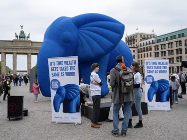 Foto van protestanten en een grote, blauwe opblaasolifant voor de Brandenburger Tor in Berlijn. Op de grond protestborden met daarop: It's time wealth gets taxed the same as work!