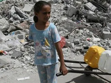 Foto van meisje in Gaza die naast een gigantische berg met puin een steekwagentje vooruittrekt met daarop jeerycans met water.