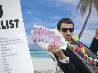 Een man in pak zit op een tropisch strand en houdt allemaal briefjes van 500 euro vast. Op de voorgrond een papier met de titel EU blacklist.