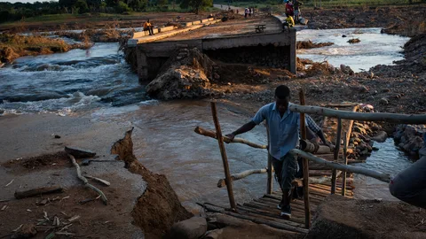 Een kapotte brug over een rivier. Op de voorgrond loopt een man de wal op.