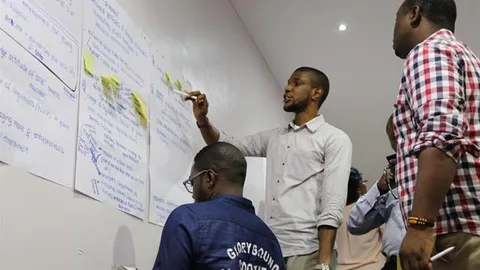 Iemand geeft les terwijl hij wijst naar papieren die hangen aan een muur.