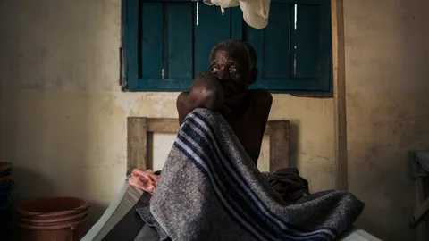 Lwako Okelwa met een deken op zijn bed in een kliniek.