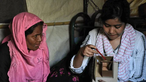 Een vrouw laat een papieren model van het toiletgebouw zien. Een andere vrouw kijkt toe.
