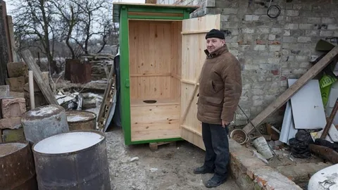 Mykola staat buiten voor een houten latrine met openstaande deur.