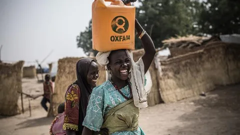 Twee vrouwen waarvan een een watertank met het Oxfam logo op haar hoofd draagt.