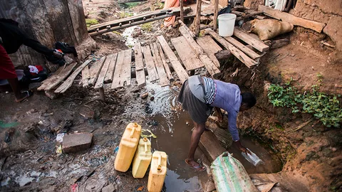 Foto van vrouw die met jerrycans water uit een gat in de straat schept.