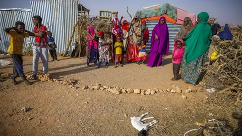 Mensen staan voor een aantal hutten. Op de voorgrond ligt een geitje.