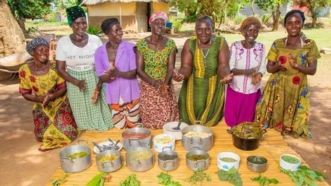 Seven women sit in line. In front of them are pans and vegetables.