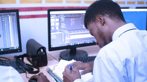 A student sits in front of a screen while writing in a notebook.