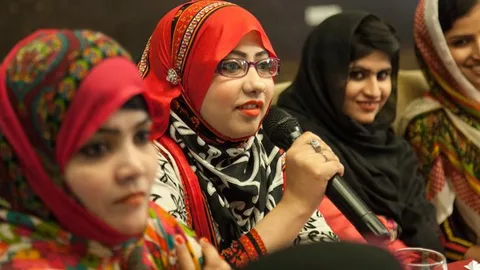 Women wearing headscarves sit next to each other. One of them speaks into a microphone.