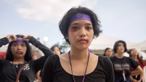 Foto van een Filipijnse vrouw met blauwe plekken in haar gezicht. tijdens een protest op vrouwendag. Ze draagt een hoofdband met de tekst "Kaisa"