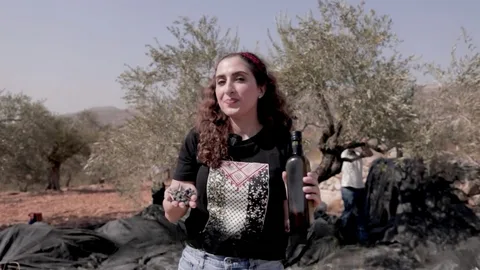 Een Palestijnse vrouw heeft olijven en een fles olijfolie in de hand, met op de achtergrond olijfbomen op de Westelijke Jordaanoever.