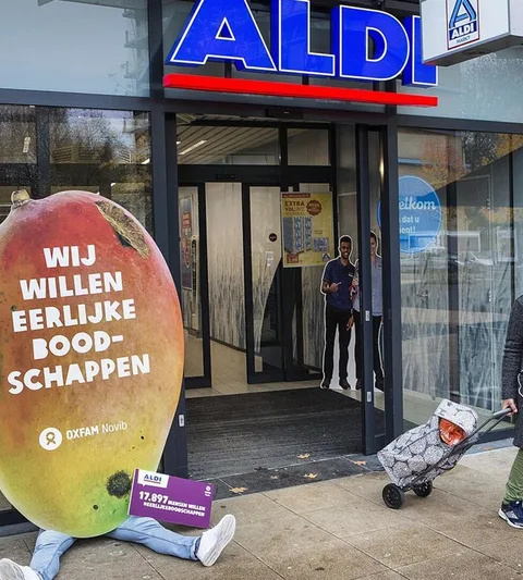 A person sits in front of a supermarket. Covering his body is a sign with the text: Wij willen eerlijke boodschappen (we want fair groceries)